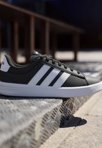 Baskets Adidas noires avec des rayures blanches reposant sur une surface métallique texturée en extérieur.