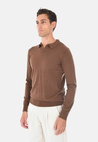 Pullover marrone intrecciato con scollo a V e colletto ripiegato; design aderente, polsini e orlo a coste; texture morbida. Abbinato a pantaloni chiari.