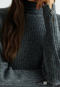 Pull en maille gris à col roulé avec texture côtelée, présentant un accent de bouton décoratif au niveau du poignet et une coupe ample.