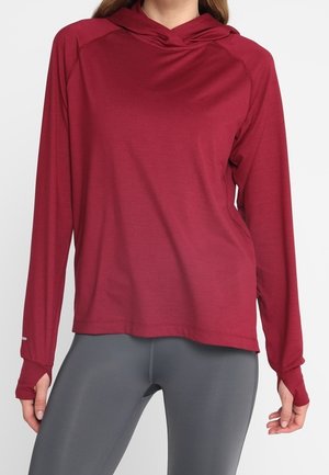 Kvinde iført en bordeaux rød hættetrøje med lange ærmer og tommelhuller samt grå tætsiddende leggings, stående mod en ensfarvet baggrund.