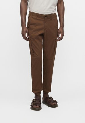 Homme portant un pantalon cargo marron avec des poignets élastiques et des sandales marron foncé à double bride, debout devant un fond uni clair.