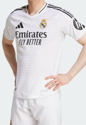 Osoba ubrana w biały strój piłkarski Realu Madryt z czarnymi paskami Adidas, logo Emirates Fly Better oraz herbem drużyny na piersi.