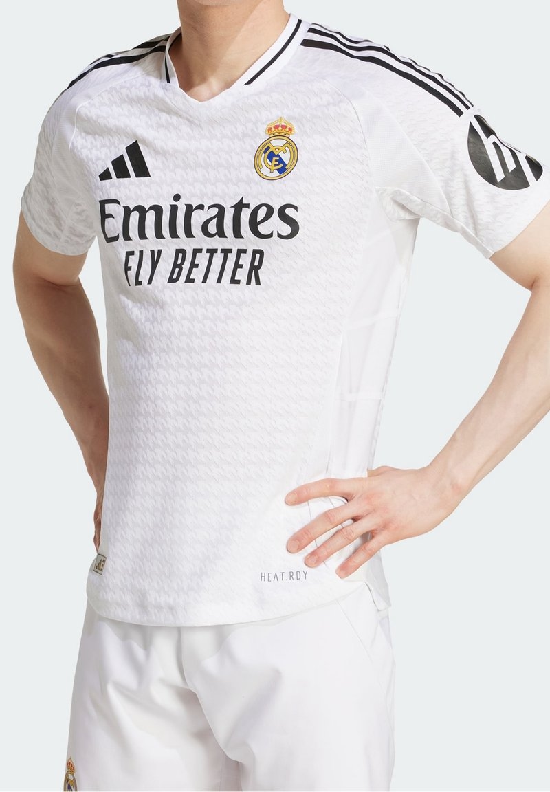 Personne portant un maillot de football blanc du Real Madrid avec des bandes noires Adidas, le logo Emirates Fly Better, et le blason de l'équipe sur la poitrine.