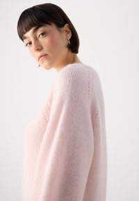 Light pink, oversized knitted sweater with a soft texture, wide sleeves, and small open stitch details near the neckline.