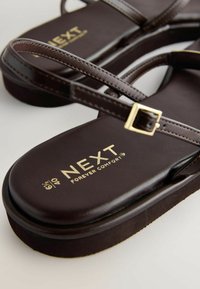 Sandales en cuir marron foncé avec de fines lanières et une petite boucle dorée, portant le logo de la marque "NEXT Forever Comfort" sur la semelle.