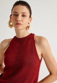 Röd stickad halterneck-top med ett texturerat mönster, öppen rygg och ribbad halsringning. Modellen bär stora krämfärgade örhängen.