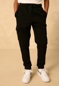 Schwarze Cargo-Hosen mit Seitentaschen, elastischem Bund und schmal zulaufenden Bündchen. Getragen mit weißen Sneakern mit schwarzen Akzenten.