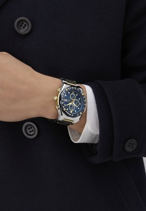 Reloj cronógrafo con una esfera azul, acentos dorados y pulsera de acero inoxidable. Las características notables incluyen tres subesferas y un bisel texturizado.