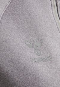 Grauer Sweatshirt mit einer weichen Textur, das ein auffälliges geprägtes Logo und Designelemente aufweist. Der Stoff hat eine melierte Optik.