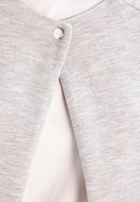 Gray knit fabric with a textured finish, featuring a snap closure and a layered design. The background is a smooth light fabric.