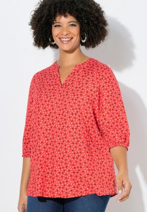 Smilende kvinde med krøllet hår iført hvide blomsterformede øreringe og en rød bluse med små hjerte-mønstre og 3/4 ærmer.