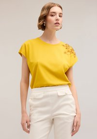 Geel T-shirt met korte mouwen met bloem borduurwerk op de schouder, gecombineerd met witte high-waisted trousers. Gladde stofstructuur.