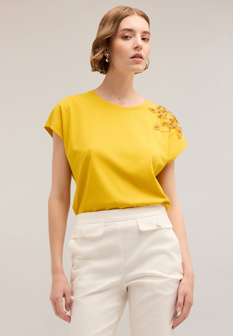 Geel T-shirt met korte mouwen met bloem borduurwerk op de schouder, gecombineerd met witte high-waisted trousers. Gladde stofstructuur.