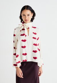Blouse crème avec un motif répétitif de lèvres rouges, dotée d'un décolleté en V et d'un nœud au cou. Tissu doux, coupe ample et manches évasées.