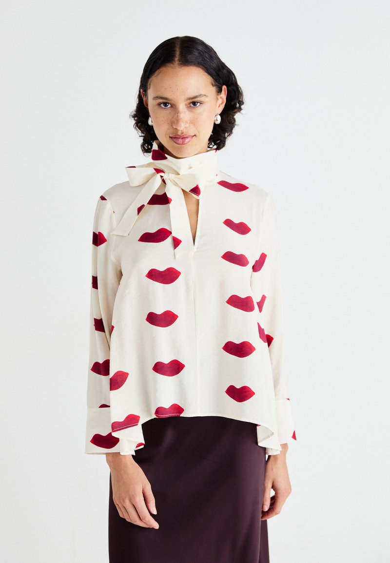 Blouse crème avec un motif répétitif de lèvres rouges, dotée d'un décolleté en V et d'un nœud au cou. Tissu doux, coupe ample et manches évasées.