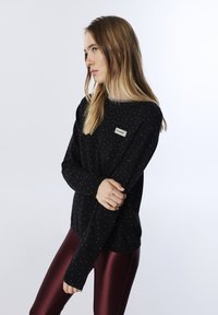 Pull noir avec des éclats blancs, col rond et manches longues, associé à des leggings bordeaux brillants. Patch notable sur le devant du pull.