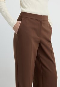 Bruine op maat gemaakte broek met een gladde textuur, voorzien van een voorzak en een platte tailleband. Gecombineerd met een aansluitende crèmekleurige geribbelde trui.
