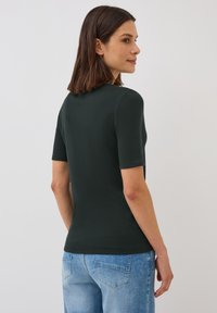 Un haut court à manches courtes de couleur vert foncé avec un col rond, une coupe ajustée et une texture lisse, associé à un jean bleu clair dans un cadre décontracté.