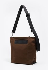 Brązowa torba na ramię z płótna z czarnym skórzanym akcentem, regulowanym czarnym paskiem, zamkiem błyskawicznym i ozdobną kieszenią z przodu.