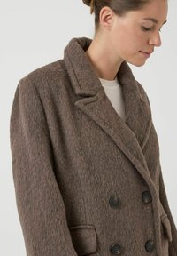 Abrigo cruzado en una tela marrón suave y texturizada. Presenta solapas anchas, dos grandes botones negros y bolsillos laterales.