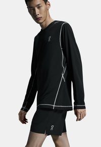 Sort langærmet atletisk top med reflekterende syninger og logo, parret med sorte shorts med sidespalter og logo ved kanten.