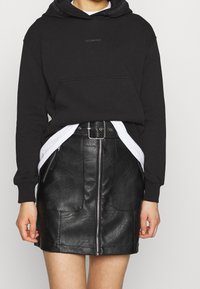 Sudadera negra con bolsillo frontal y logo en el cuello, combinada con una minifalda de cuero negra con cremallera frontal y bolsillos laterales.