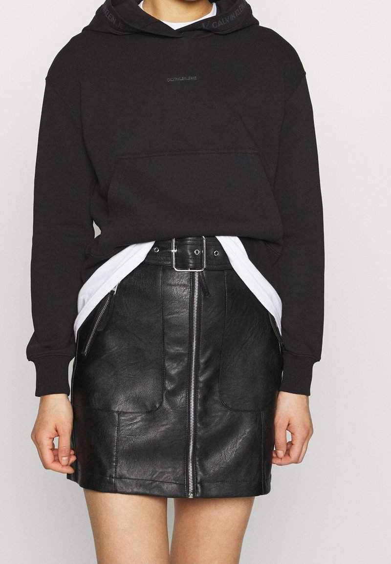 Sudadera negra con bolsillo frontal y logo en el cuello, combinada con una minifalda de cuero negra con cremallera frontal y bolsillos laterales.