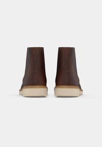 Une paire de bottines en cuir marron avec des semelles en caoutchouc beiges, vue de l'arrière sur fond blanc uni.