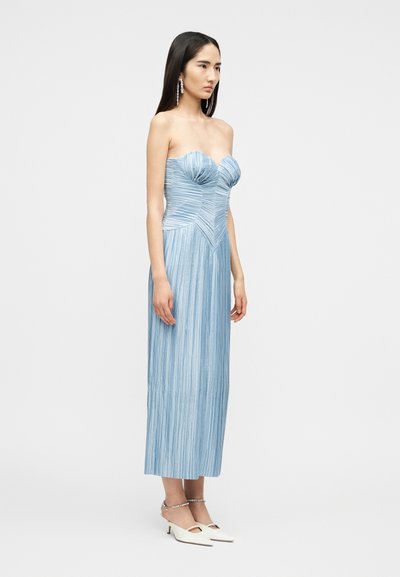 Γυναίκα που στέκεται φορώντας ένα μπλε ανοιχτό, μίντι φόρεμα χωρίς τιράντες με πτυχώσεις, εφαρμοστό μπούστο και λευκά παπούτσια με χαμηλό τακούνι και λουράκια στον αστράγαλο.