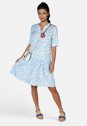 Hellblaues Kleid mit weißem Wellenmuster, kurzen Ärmeln, V-Ausschnitt und gerafftem Rock. Kombiniert mit blauen Sandalen und einer weißen Handtasche mit rotem Accessoire.