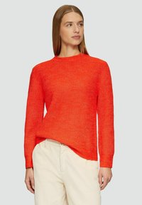 Helle orange gestrickte Pullover mit strukturiertem Muster, rundem Ausschnitt und langen Ärmeln. Kombiniert mit hellen Hosen.