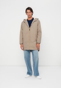 Beige gewatteerde jas met een capuchon, voorzien van een voorkruis met gele, witte en marineblauwe strepen. Gedragen over een blauwe top en lichtblauwe jeans.