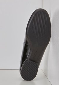 Černá lakovaná kožená bota s texturovanou gumovou podrážkou, která má liniové vzorování a vyražené logo značky. Design s zaoblenou špičkou.