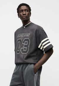 Tamnosiva Adidas majica od kordurovog materijala s velikim brojem "03" i logotipom, s bijelim prugastim rukavima i visokim ovratnikom, uparena sa sivim trenirkim hlačama.