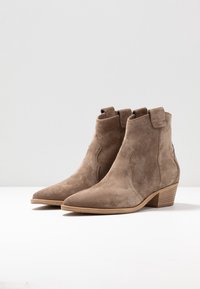 Botas de tobillo de ante marrón con punta puntiaguda, costuras decorativas y un tacón de madera apilada. Cuenta con tiradores en los lados.