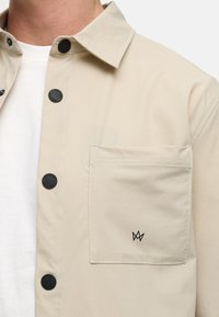 Beige skjorte med knappestolpe laget av glatt stoff, med svarte trykknapper og en brystlomme med en liten svart logo.