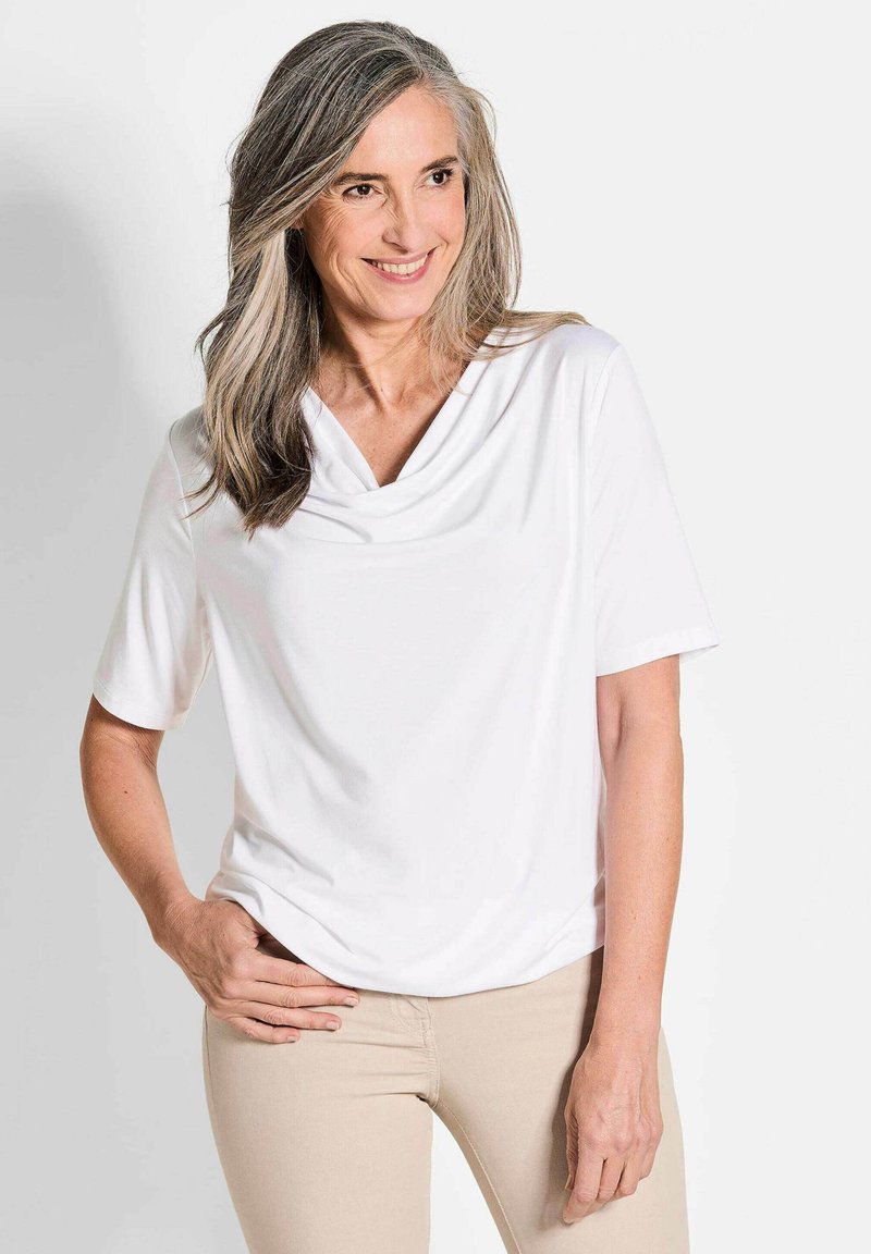 Witte top met korte mouwen en een gedrapeerde halslijn, met een gladde textuur. Het model draagt deze met beige aansluitende broek, wat een ontspannen pasvorm laat zien.