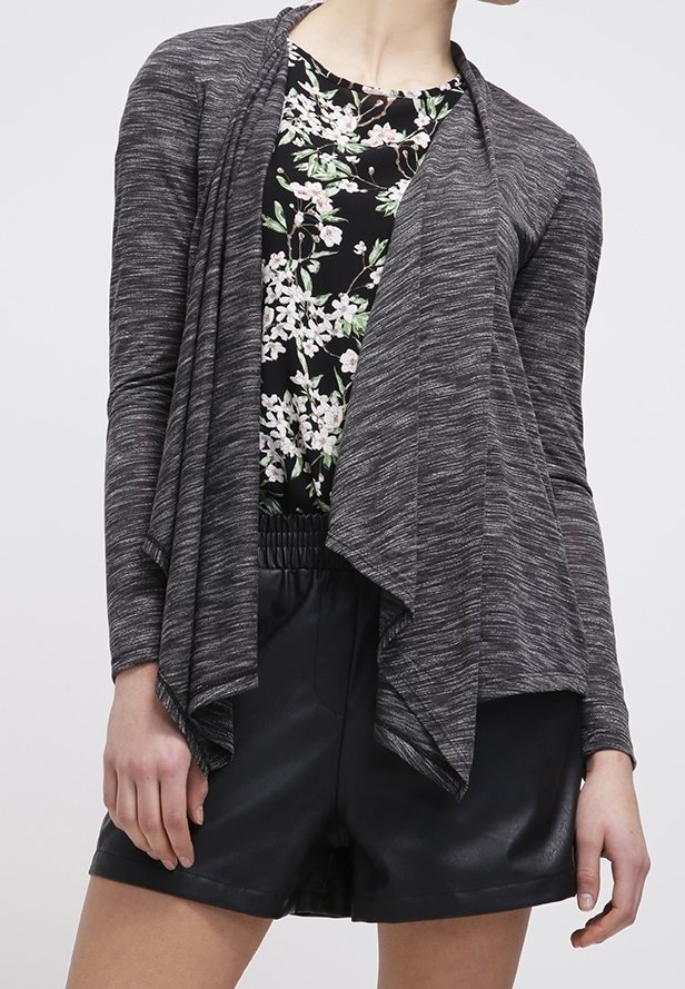 Femme portant un haut noir à motifs floraux sous un cardigan gris foncé drapé, associé à un short taille haute en cuir noir.