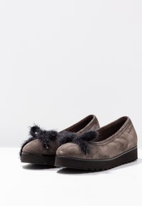 Zapatos de ante gris tipo slip-on con un acento de pompón negro texturizado en la parte delantera, suela gruesa negra y diseño de punta redonda.