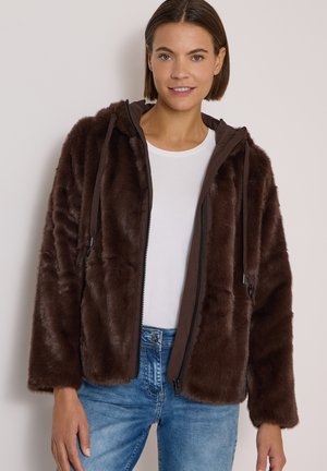 Femme portant une veste zippée en fausse fourrure marron avec une capuche, un haut blanc et un jean bleu, debout devant un fond uni clair.