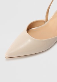Chaussure à talon slingback en cuir verni beige avec un bout pointu, une texture lisse et une finition subtilement brillante, dotée d'une fine sangle autour de la cheville.