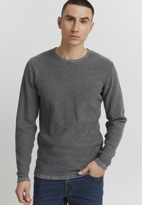 Maglietta grigia a maniche lunghe in maglia strutturata, con scollo rotondo e vestibilità casual, abbinata a jeans blu scuro.