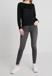 Felpa nera con logo "AX" in rilievo, abbinata a jeans skinny grigi scuri a vita alta e sneakers bianche. Design semplice e moderno.