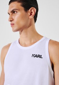 Top smanicato bianco realizzato in tessuto liscio, con il logo nero "KARL" sul lato sinistro. Design semplice con scollatura arrotondata.