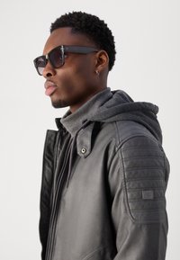 Schwarze Lederjacke mit gesteppten Schulterakzenten, über einem grauen Hoodie getragen. Das Model trägt dunkle Sonnenbrillen und hat kurzes, strukturiertes Haar.