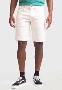 Beige Denim-Shorts mit geradem Schnitt, ausgestattet mit Fronttaschen und einem klassischen Knopfverschluss. Getragen mit schwarzen Sneakers und gemusterten Socken.