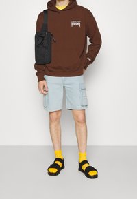 Hnědá mikina s kapucí a bílým textem, světle modré cargo šortky a černé sandály na žluté ponožky. Obsahuje černou krosnu.