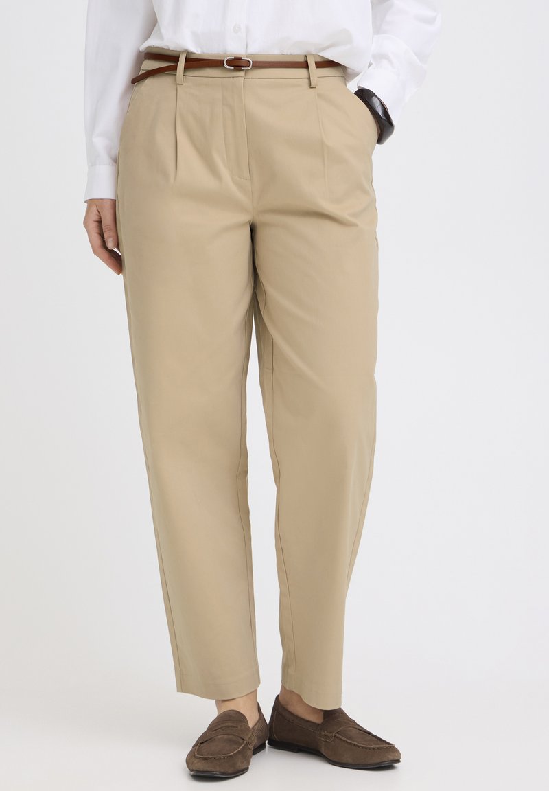 Beige maßgeschneiderte Hose mit vorderen Falten, kombiniert mit einem braunen Gürtel, weißem Hemd, schwarzer Armbanduhr und braunen Wildleder-Loafers.