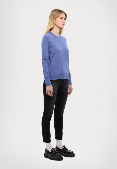 Pull bleu à manches longues avec un col rond, associé à un jean skinny noir et des chaussures à plateforme noires. Le modèle se tient de côté.