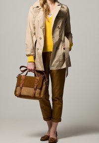 Beige trenchcoat över en gul tröja, bruna capribyxor och bruna loafers. Bär en brun och tanfärgad axelväska med spänne.